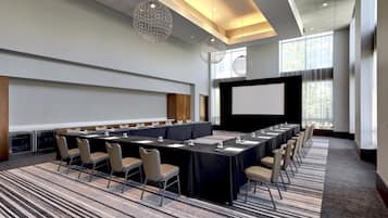 Un salón de conferencias con una larga mesa, sillas, una pantalla de proyector y lámparas colgantes.