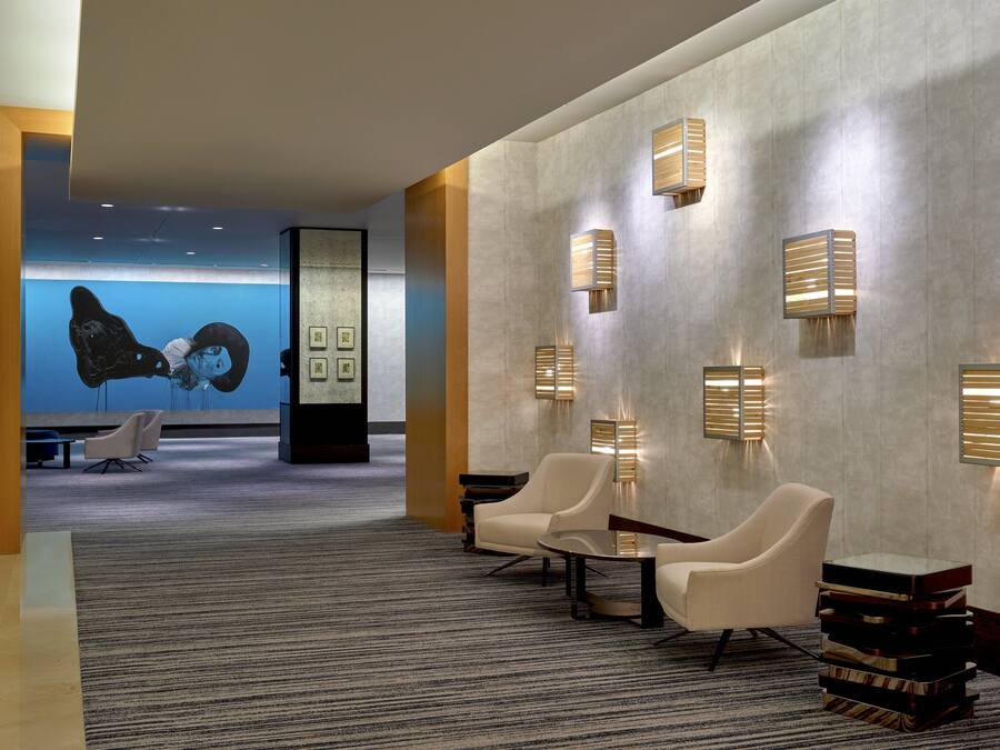 A modern lobby with a carpeted floor, a seating area with chairs and a table, and wall-mounted lighting fixtures.