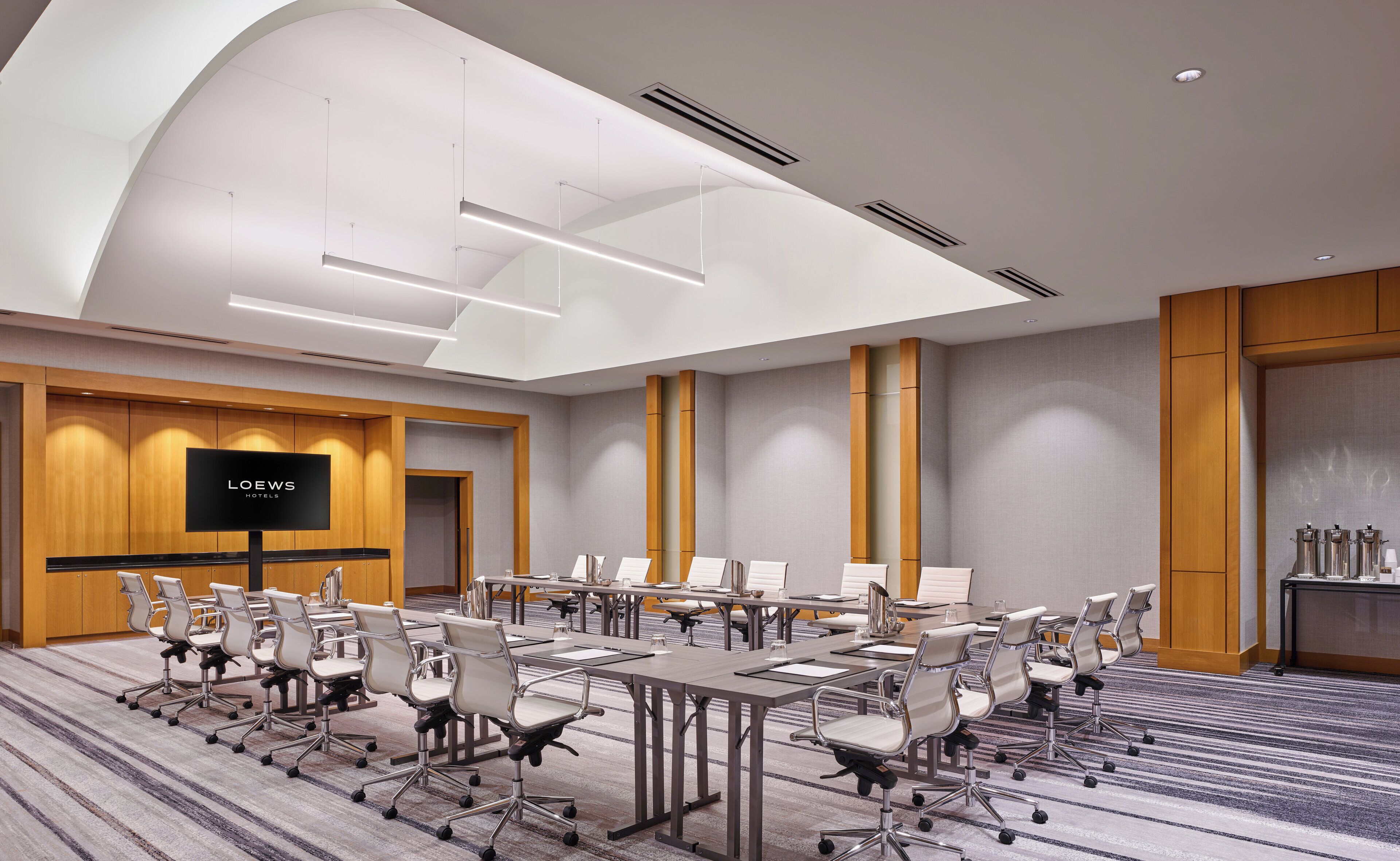 Uma sala de conferências com várias mesas e cadeiras, uma tela grande exibindo "LOEW'S HOTELS" e um design de teto moderno.