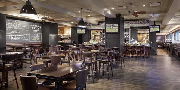 A restaurant with multiple tables and chairs, a bar area, and two televisions mounted on the wall. - Loews Chicago O'Hare Hotel (Rosemont)