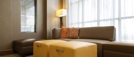 A hotel room with a sofa, two ottomans, a lamp, and a window with blinds.