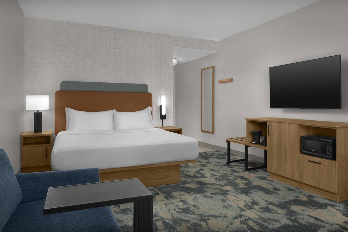 Ein Hotelzimmer mit Bett, Fernseher, Mikrowelle, Kaffeemaschine, Schreibtisch und Stuhl.