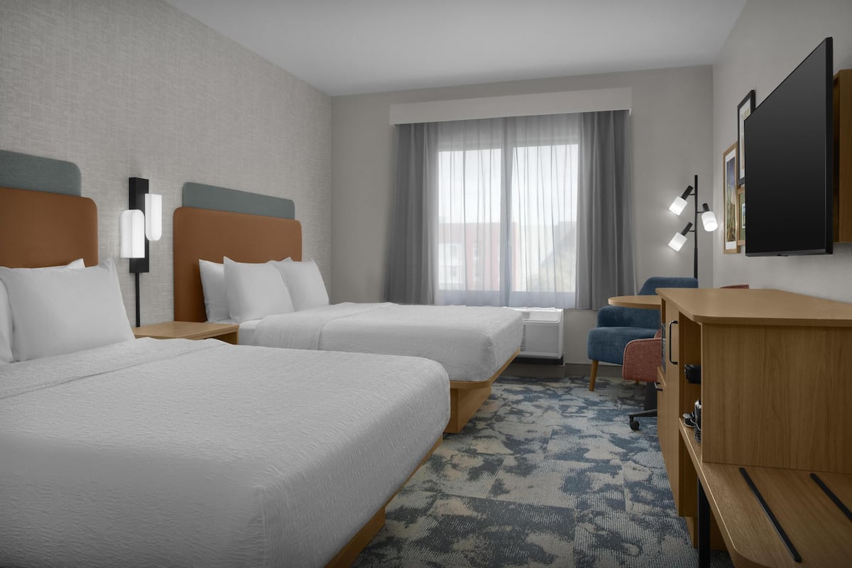 Ein Hotelzimmer mit zwei Betten, einem Schreibtisch, einem Stuhl, einem Fernseher und einem Fenster mit Vorhängen.