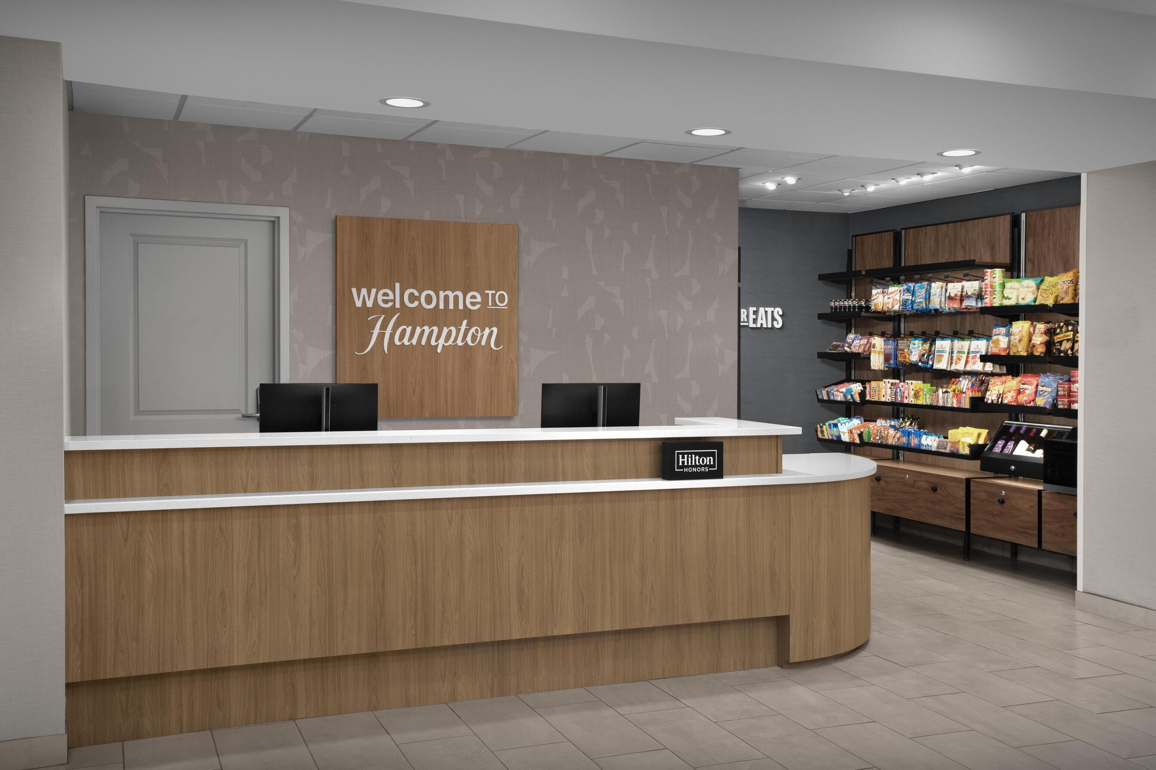 A hotel reception desk with a 'Welcome to Hampton' sign, a minibar, and snack shelves.