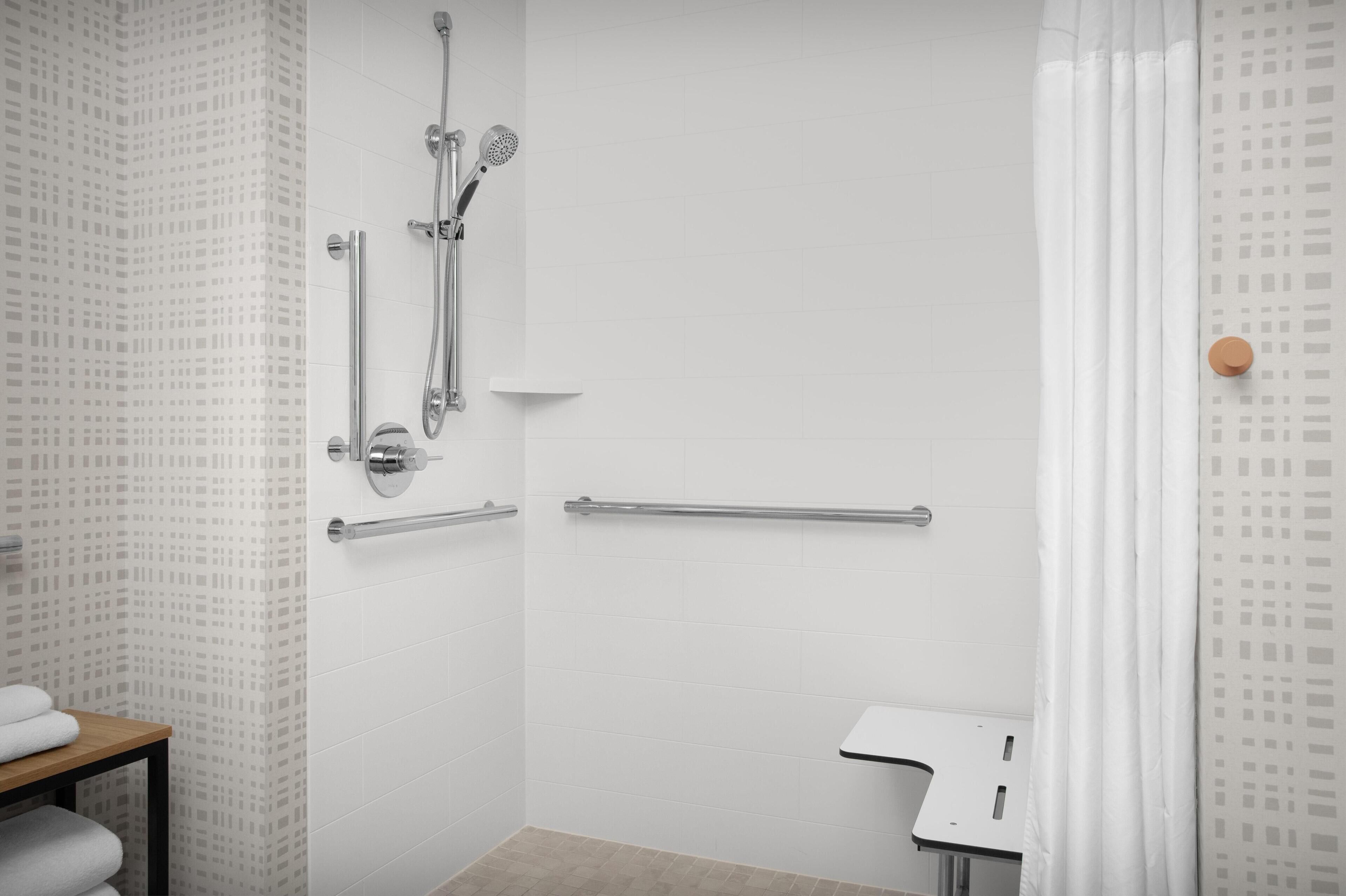A modern bathroom with white tiled walls, a shower with a handheld showerhead, a grab bar, and a toilet with a white seat.