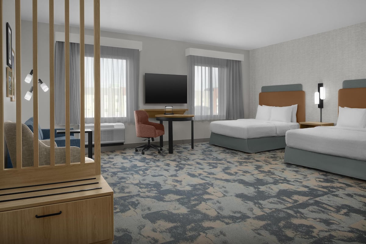 Ein Hotelzimmer mit zwei Betten, einem Fernseher, einem Schreibtisch, einem Sessel und einem Babybett.