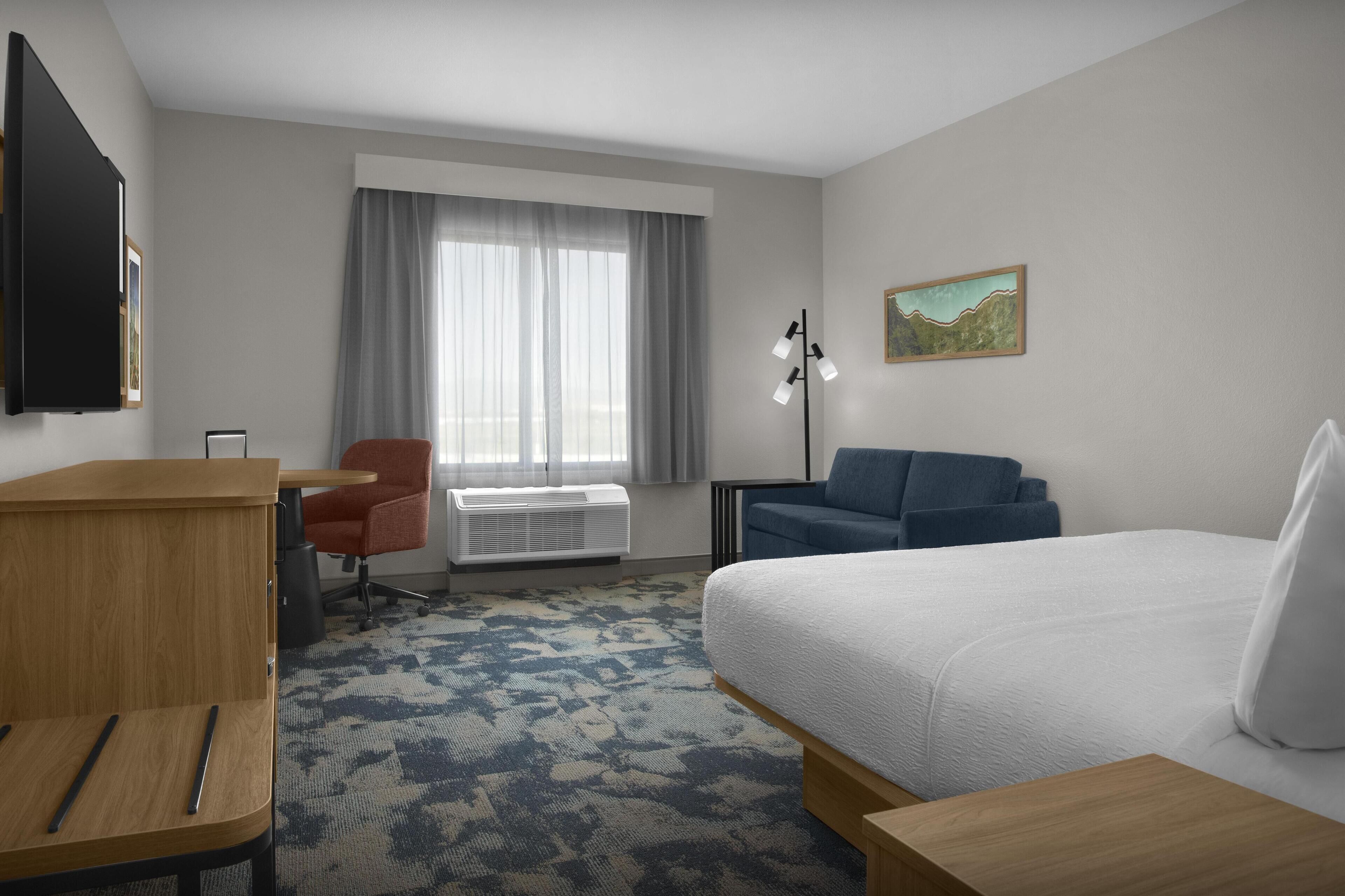 A hotel room with a bed, a desk, a chair, a standing lamp, and a window with curtains.