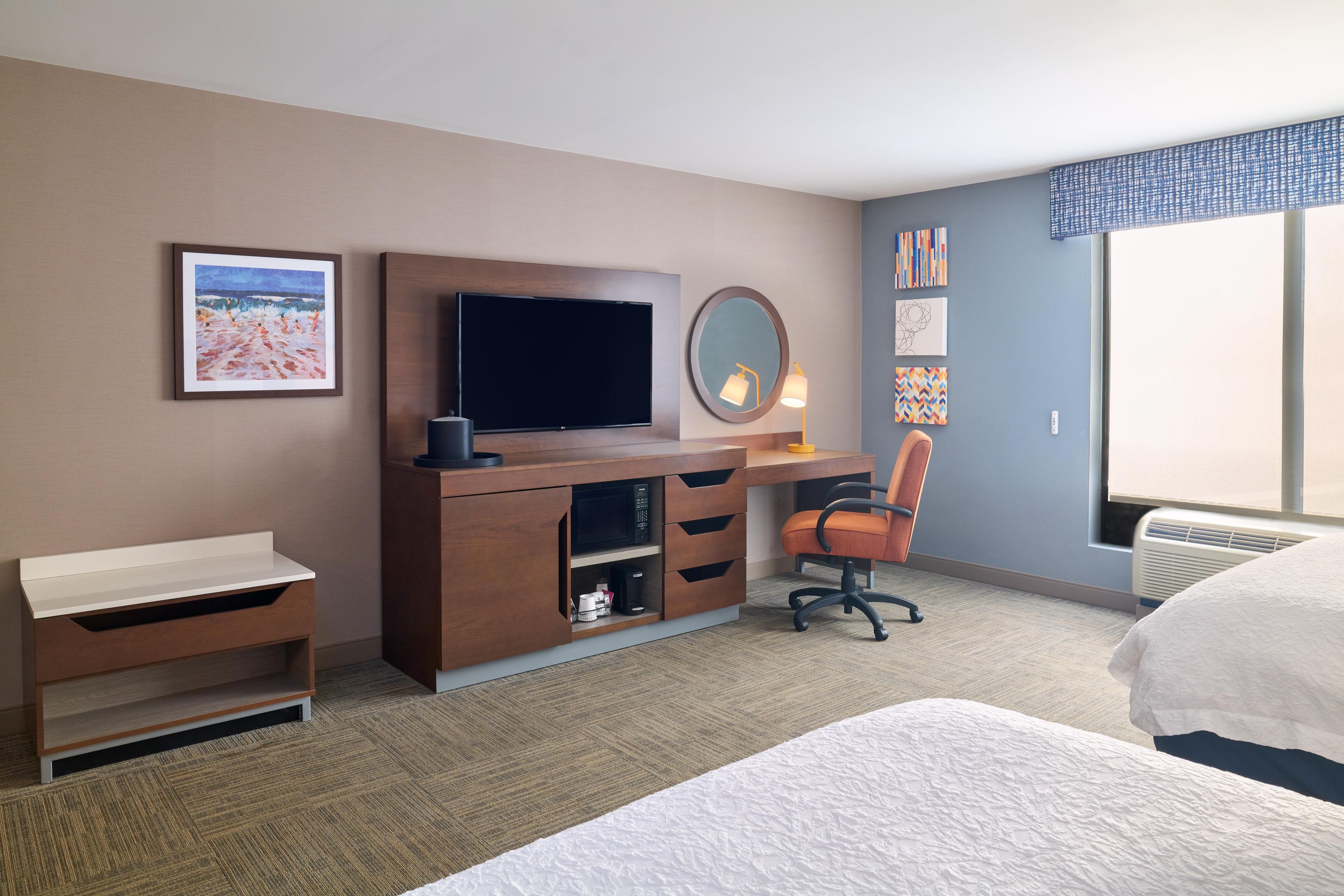 A hotel room with a bed, a desk with a flat-screen TV, a chair, a dresser, a mirror, and artwork on the wall.
