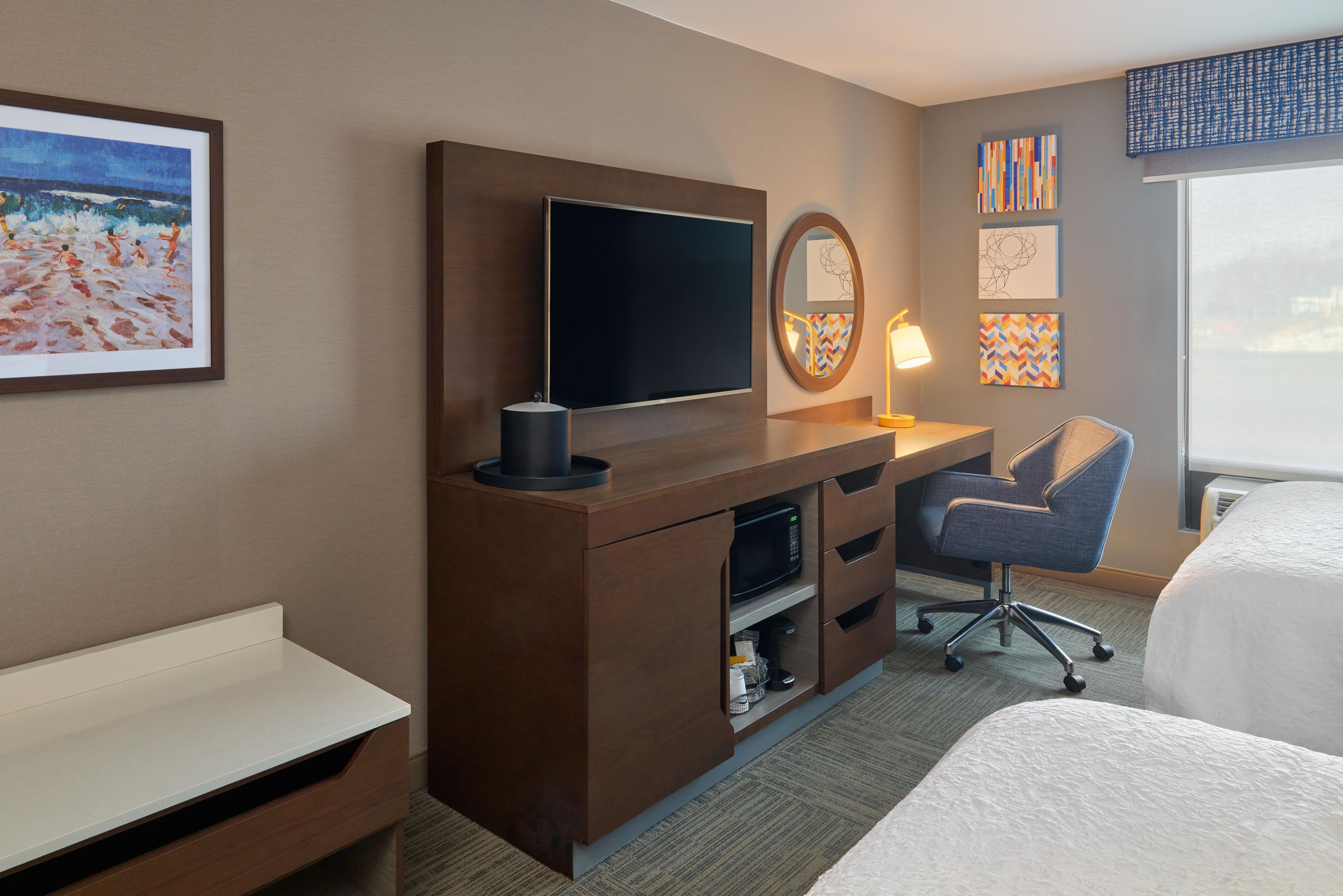 A hotel room with a bed, a desk with a flat-screen TV, a chair, a lamp, and artwork on the walls.