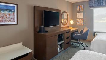 A hotel room with a bed, a desk with a flat-screen TV, a chair, a lamp, and artwork on the walls.