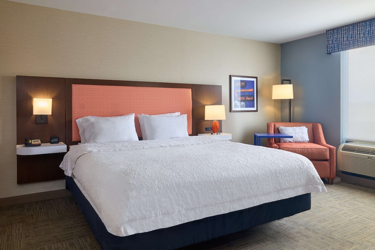 Quarto de hotel com uma cama grande, duas mesas de cabeceira com abajures, uma cadeira e um quadro na parede.