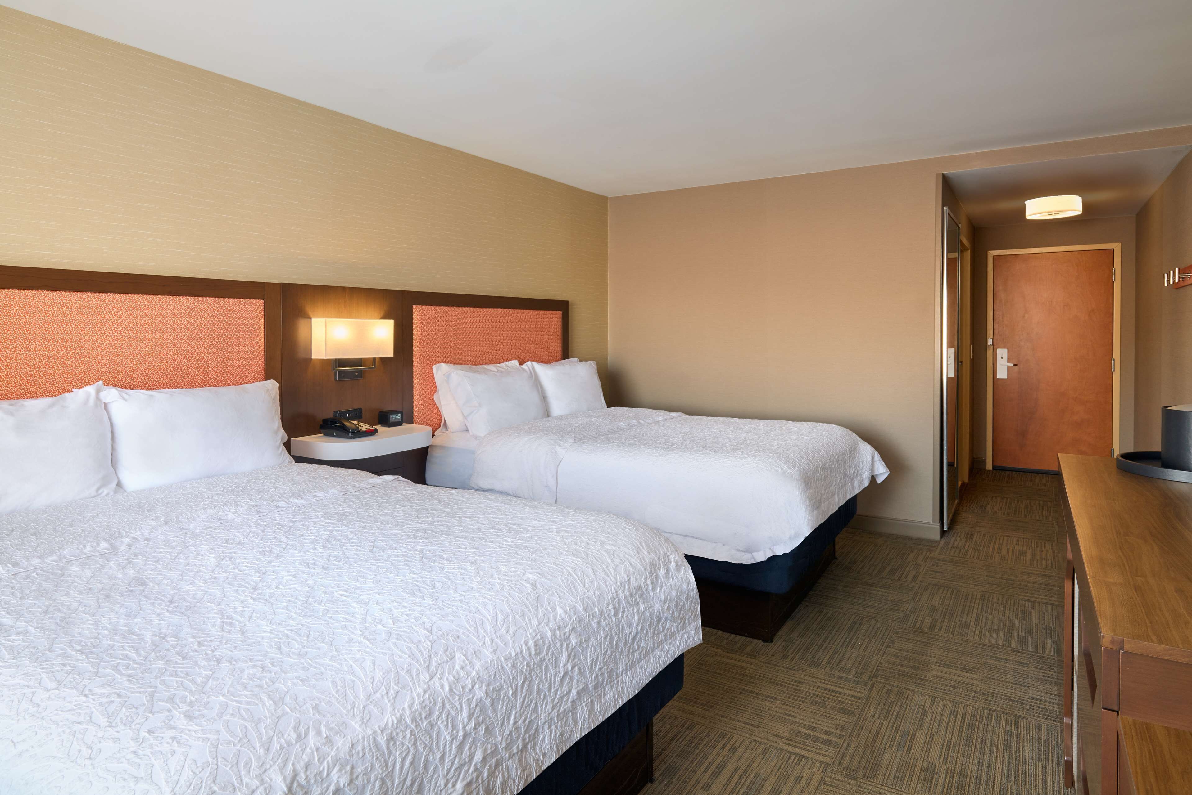 A hotel room with two beds, a nightstand, a lamp, and a wooden desk.
