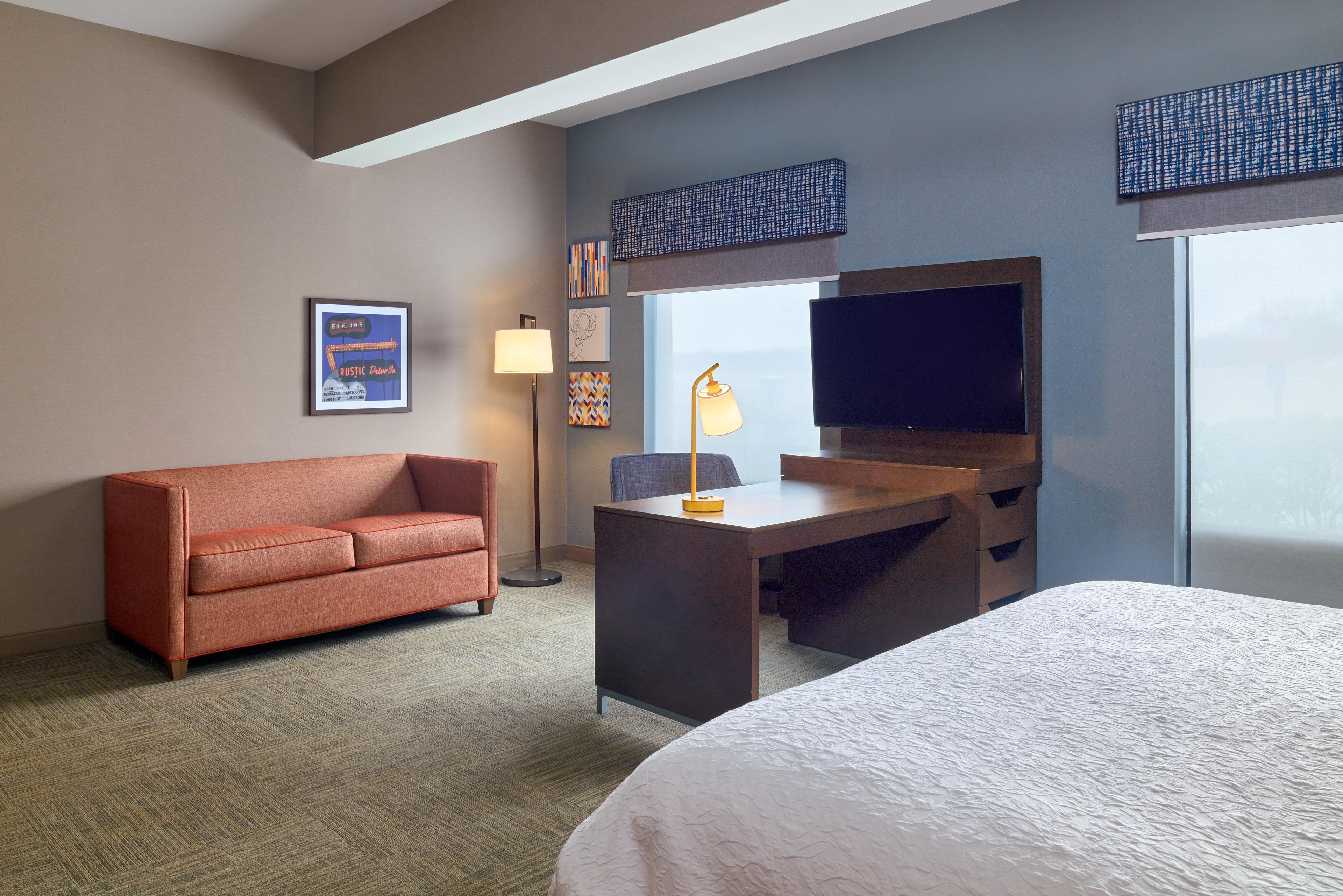 A hotel room with a bed, a desk, a sofa, a TV, and artwork on the wall.