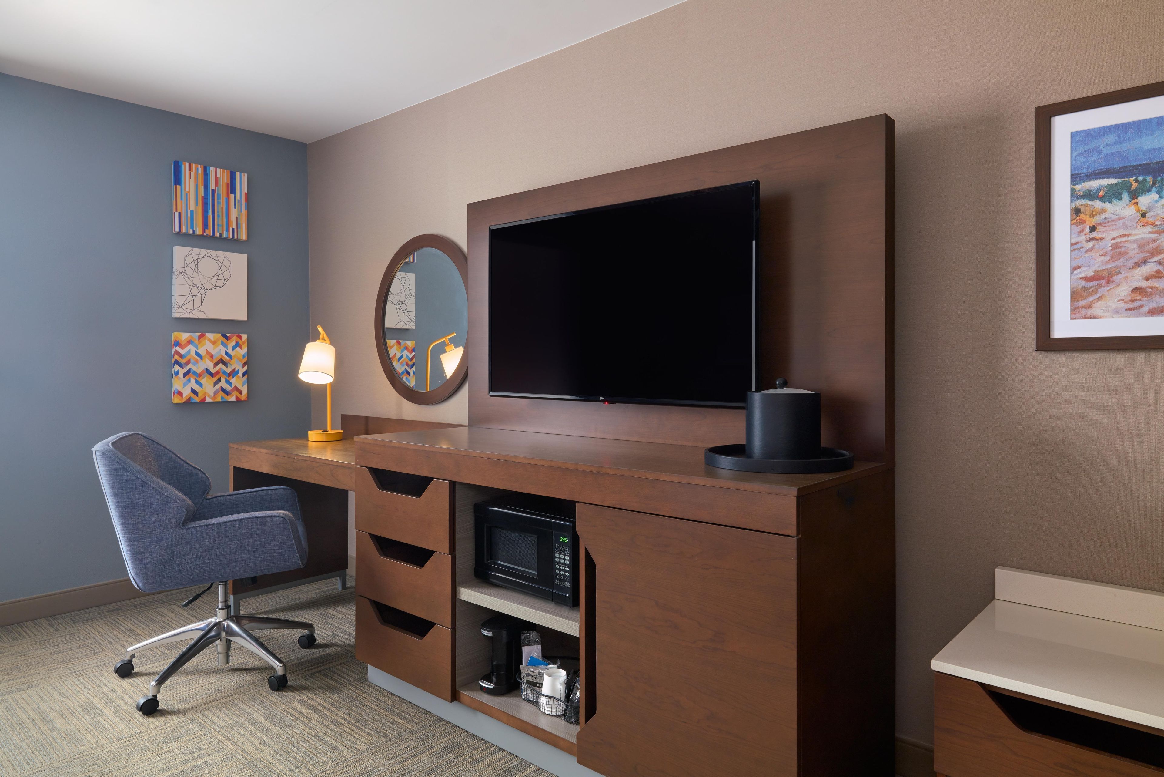 Une chambre d’hôtel moderne avec un bureau en bois, une chaise, un téléviseur à écran plat, un miroir et des œuvres d’art accrochées aux murs.