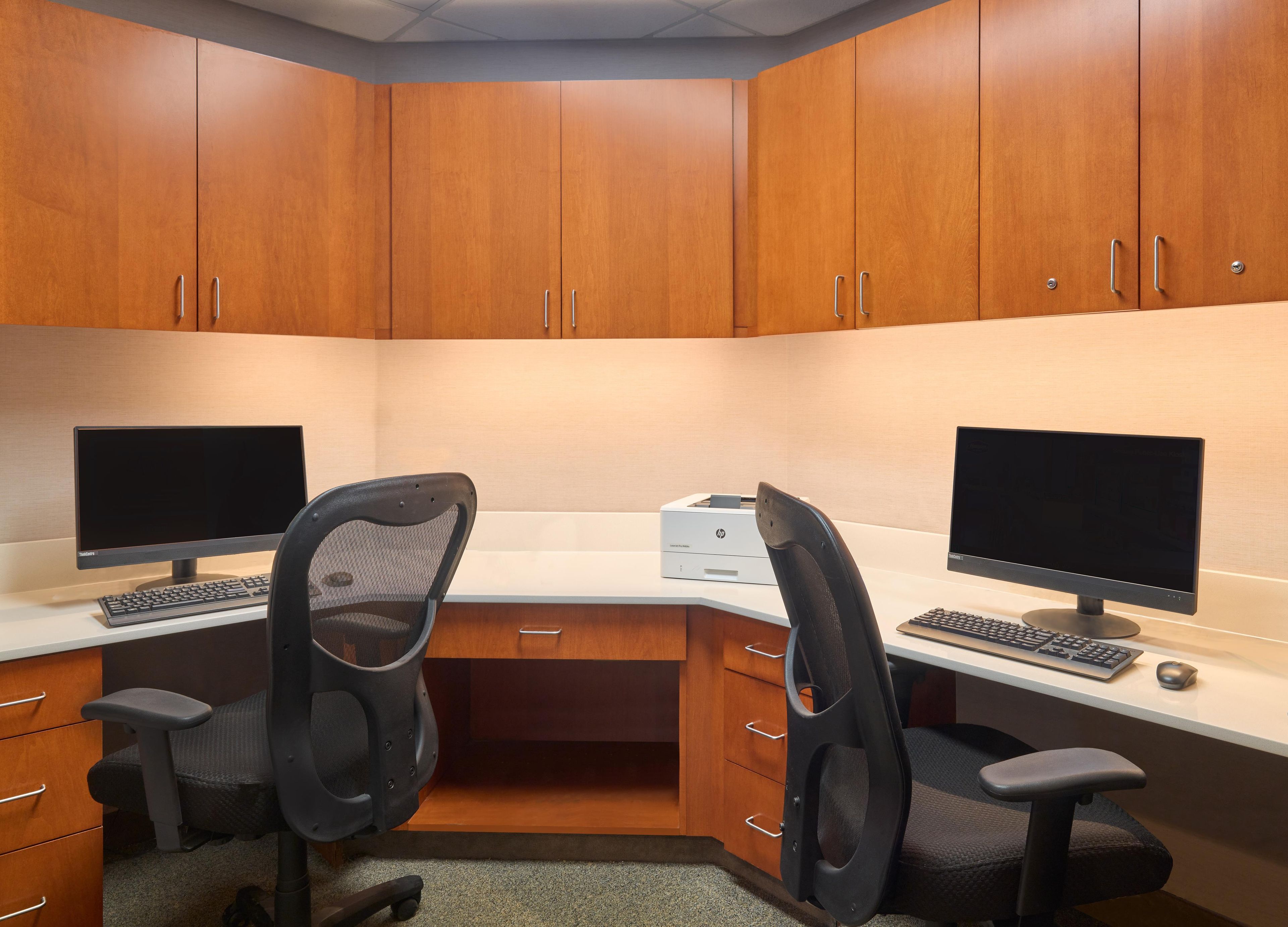 A corner office with two desks, each with a computer monitor, keyboard, and chair. There is a printer on the desk, and the room has wooden cabinets.