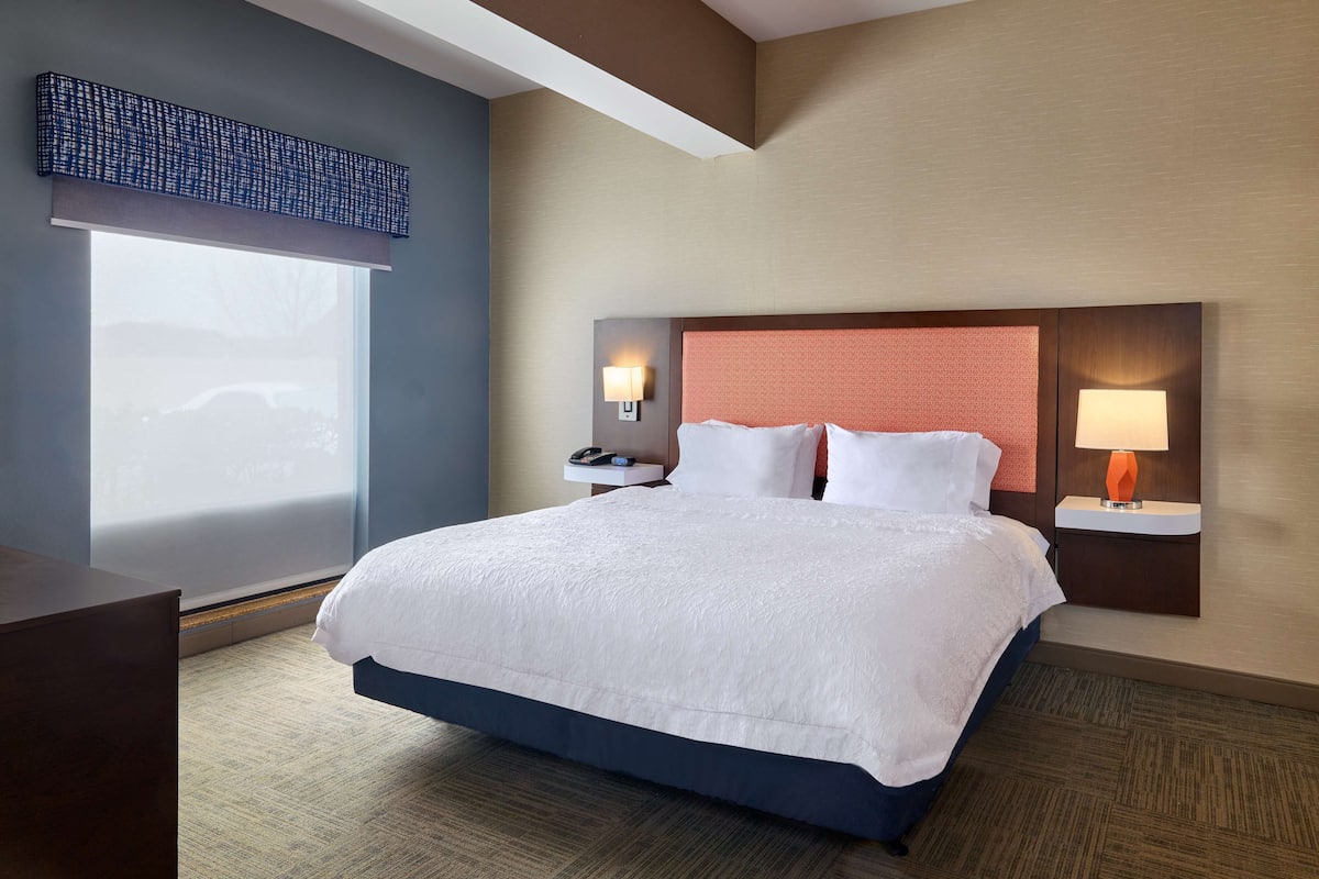 Quarto de hotel com uma cama grande, duas mesas de cabeceira com luminárias, uma mesa de apoio e uma janela com vista para um carro.