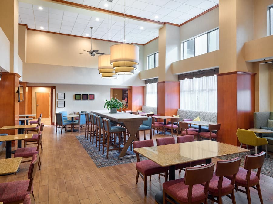 Un hall d’hôtel spacieux, doté de plusieurs espaces de détente, notamment une grande table à manger centrale entourée de chaises, et un coin salon avec vue sur l’extérieur.