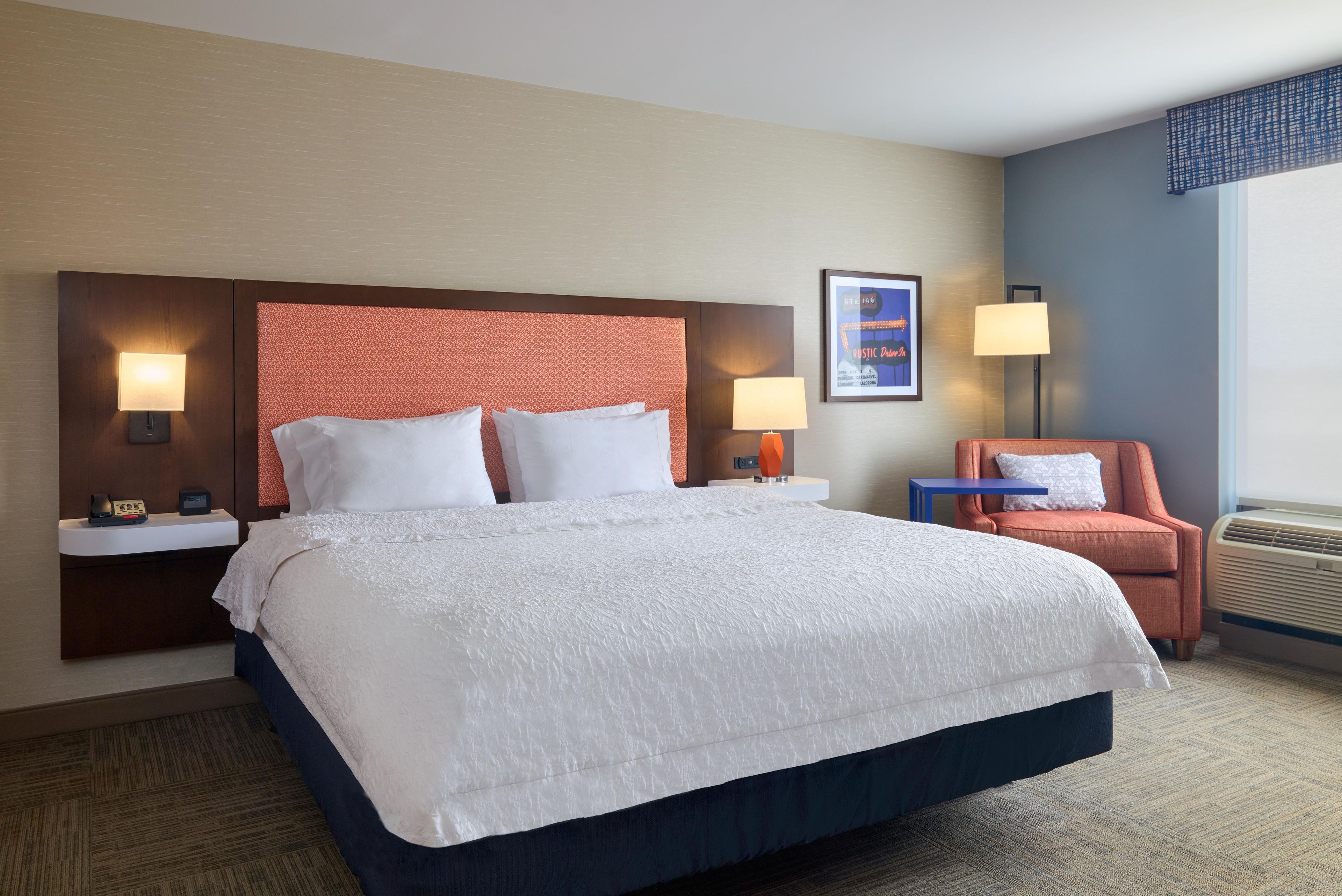 A hotel room with a large bed, two bedside tables with lamps, a small table, a chair, and a wall-mounted air conditioner.