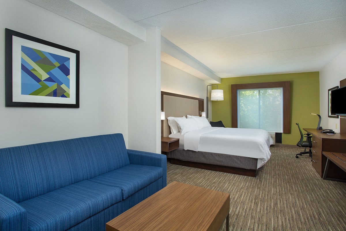 A hotel room with a blue sofa, a bed with white bedding, a desk with a computer, and a framed abstract artwork on the wall.