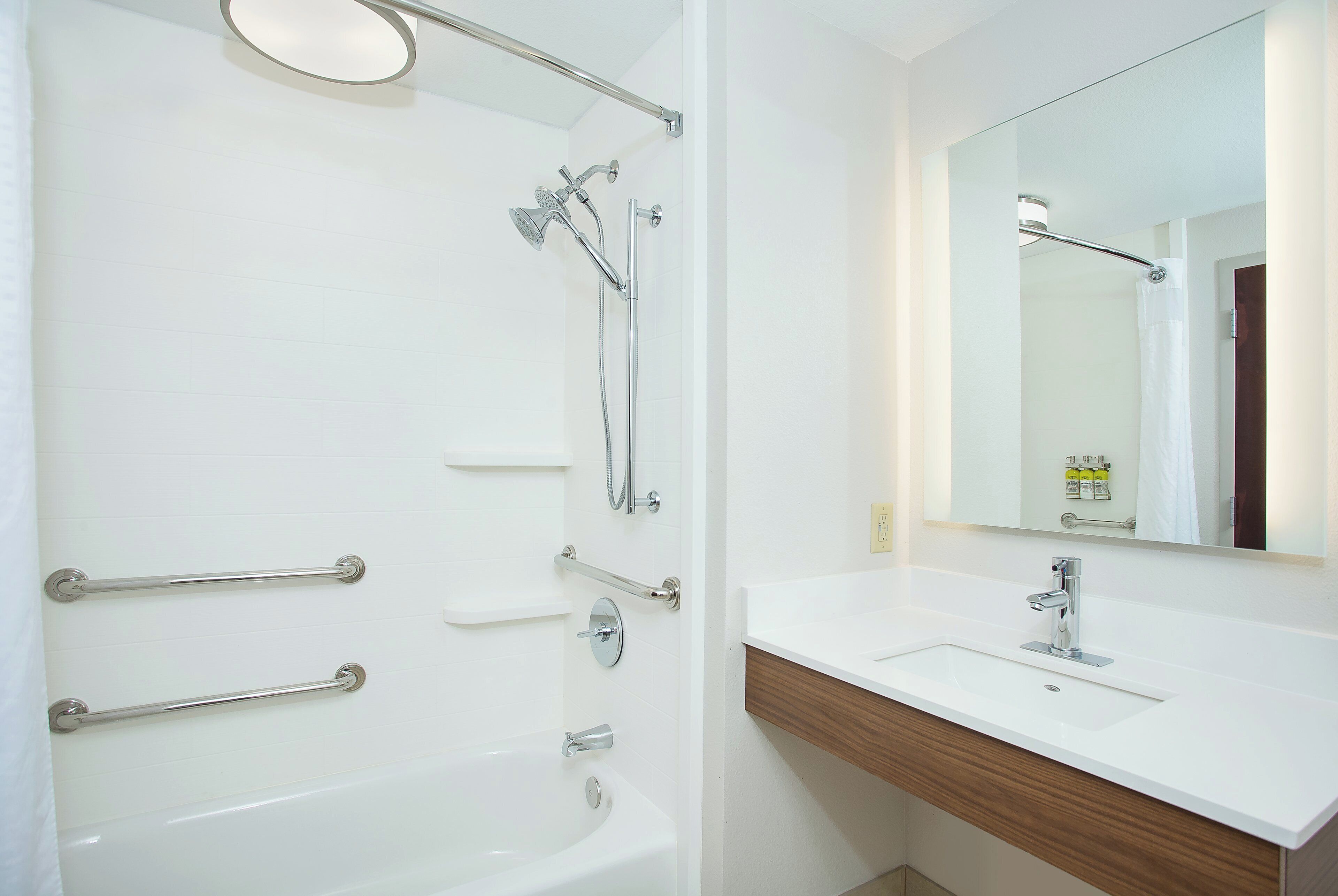 A modern bathroom with a bathtub, a shower, a vanity with a mirror, and handrails.