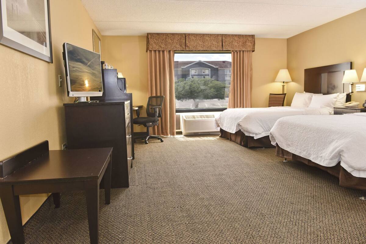 A hotel room with two beds, a desk, a chair, a television, and a window with a view of buildings.
