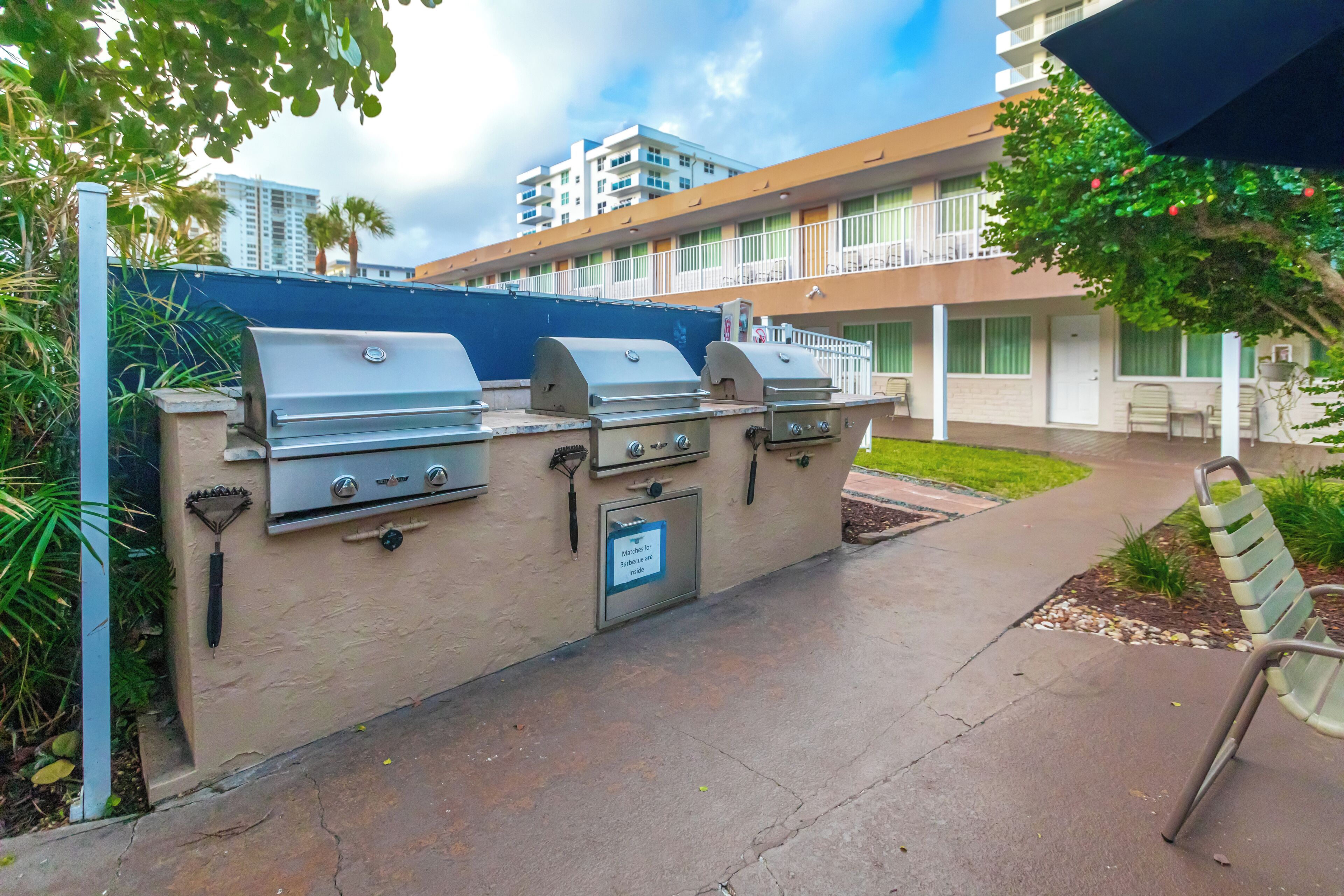 An outdoor barbecue area with multiple grills, a sign indicating matches for barbecue, and a building with balconies in the background.