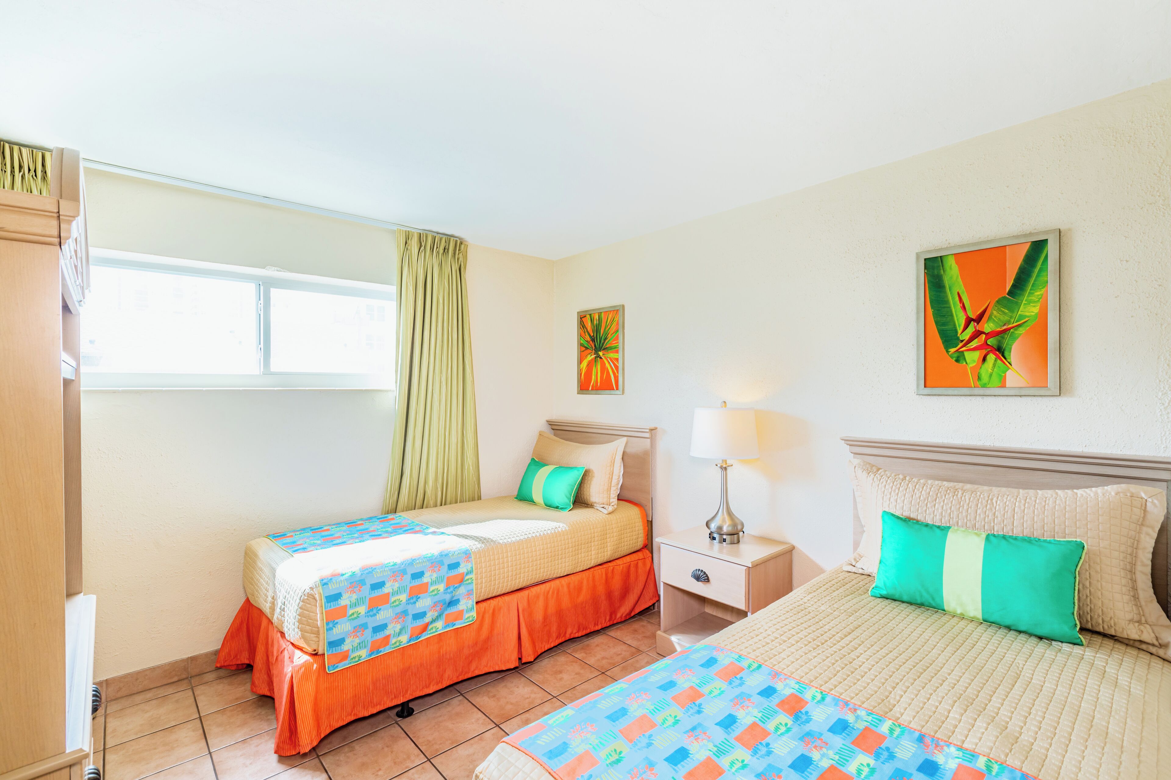 Une chambre d’hôtel avec deux lits, une fenêtre avec des rideaux, une table de chevet avec une lampe et des œuvres d’art accrochées aux murs.