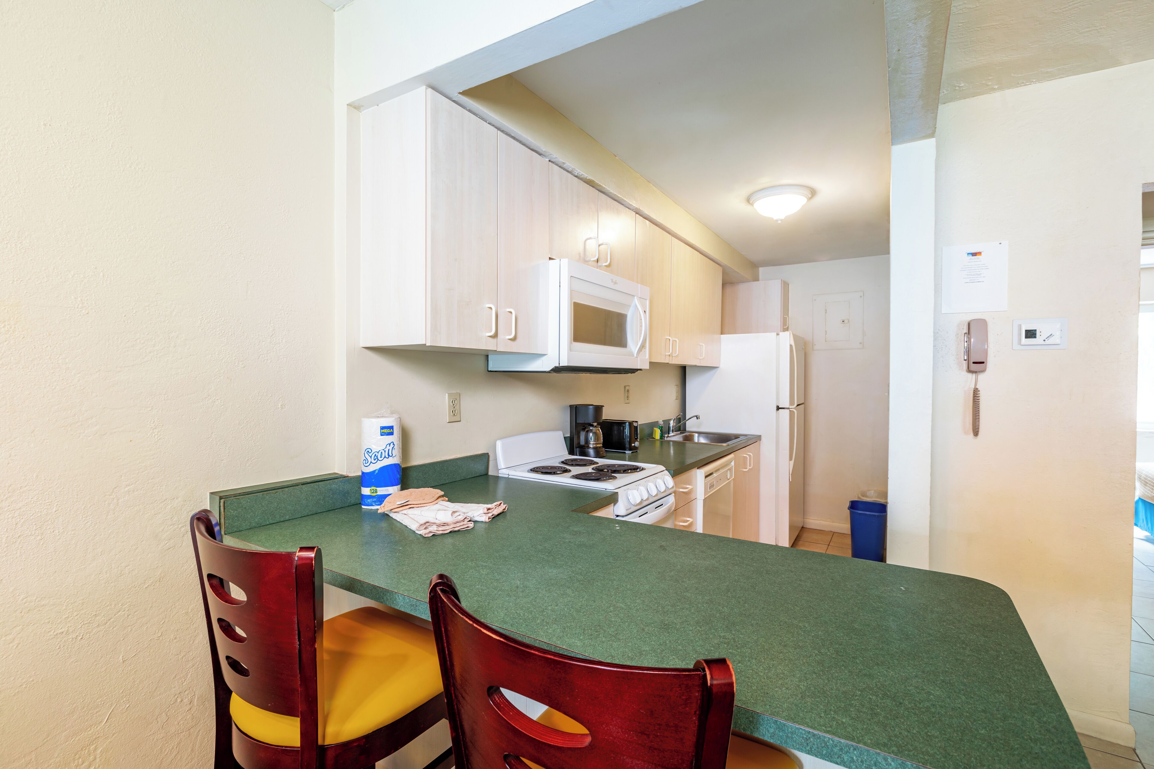 A compact kitchen with a green countertop, white cabinets, a stove, and a microwave. There are two wooden chairs with yellow cushions.