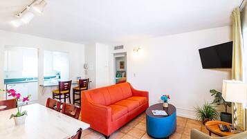 Uma sala de estar com um sofá vermelho, uma mesa de jantar com cadeiras, uma mesinha lateral azul e uma TV fixada na parede.
