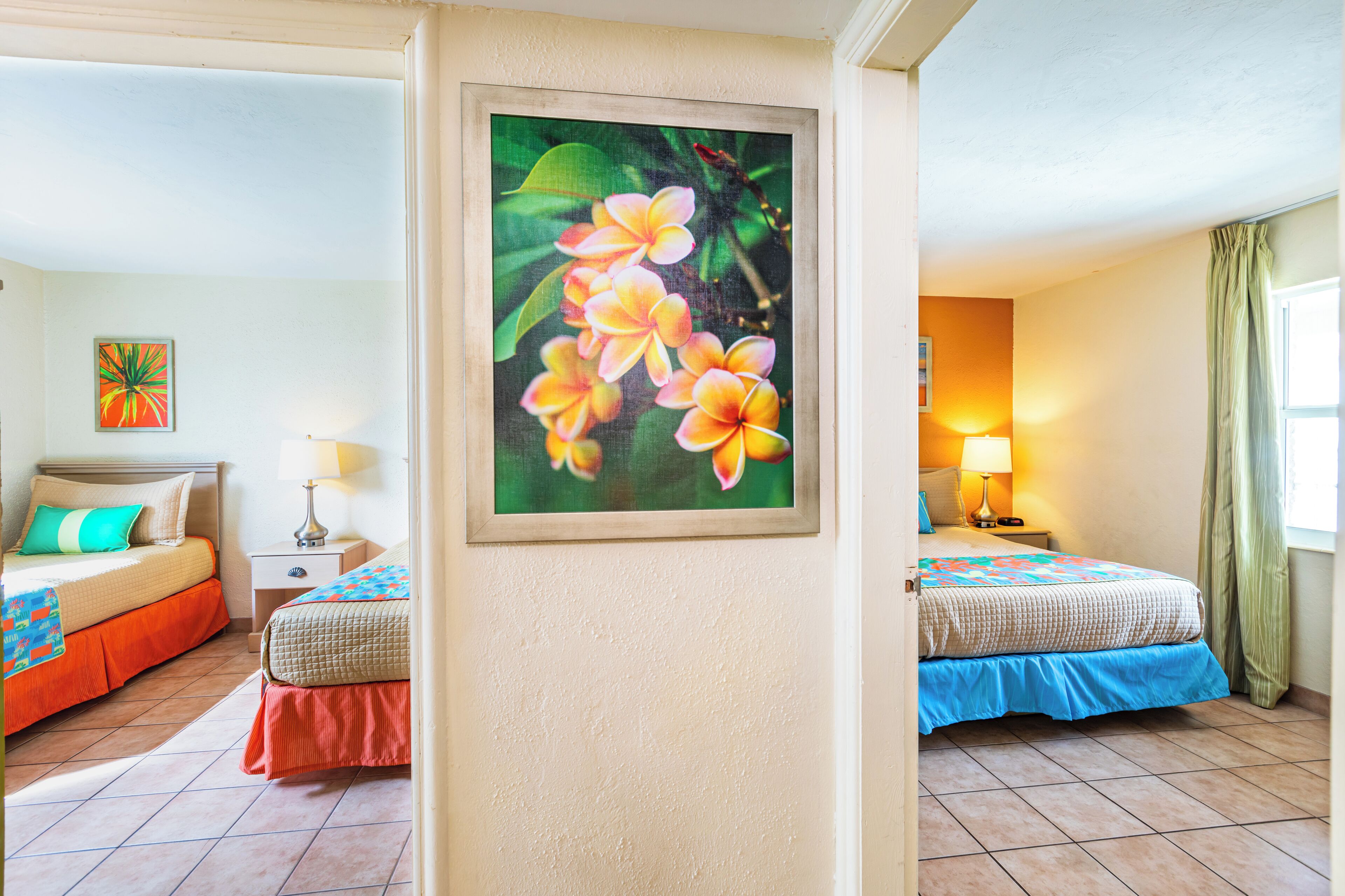 A hotel room with two beds, a nightstand, a lamp, a painting of flowers, and a window with curtains.