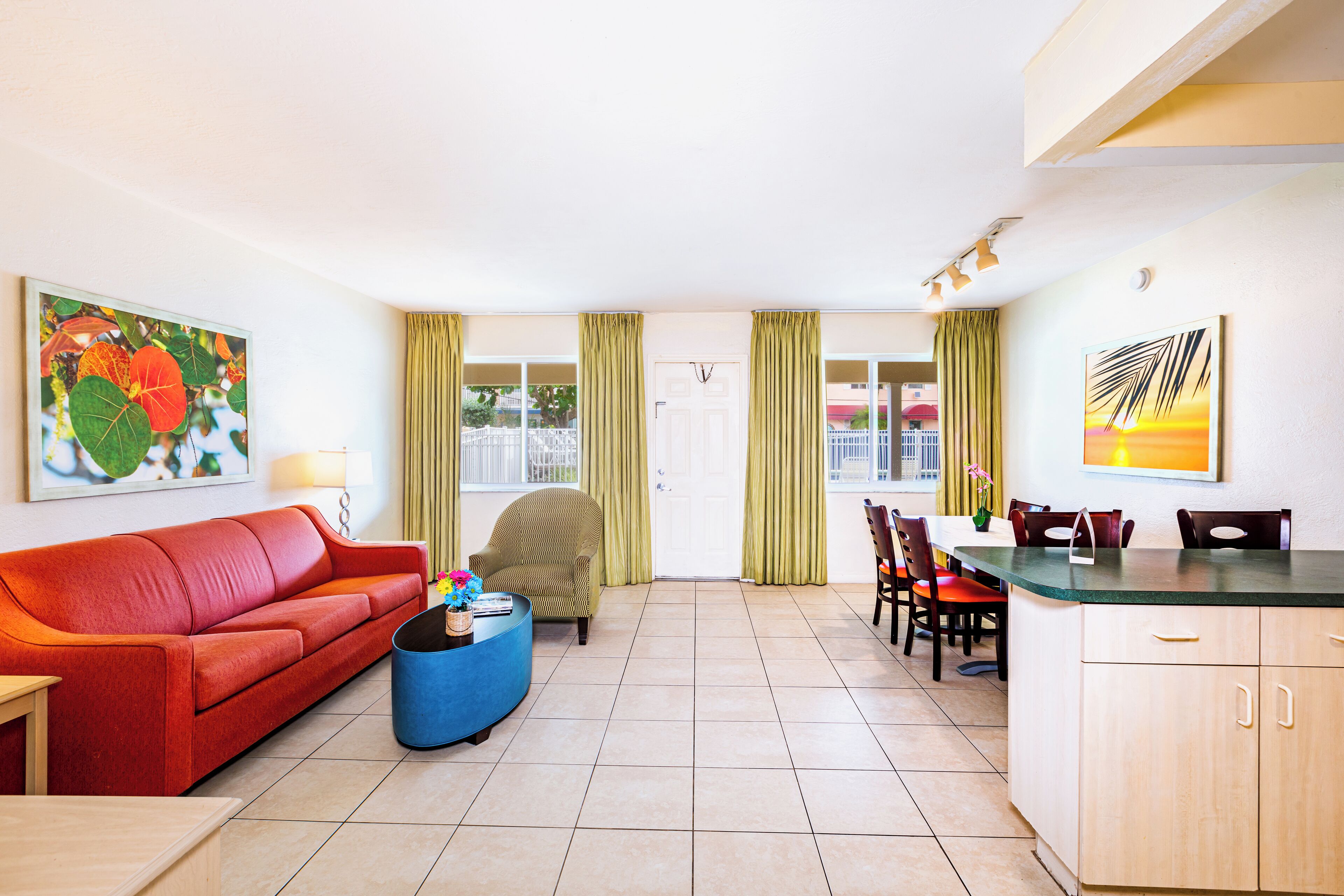 Un salon avec un canapé rouge, un pouf bleu, un fauteuil vert et un coin repas comprenant une table et des chaises.