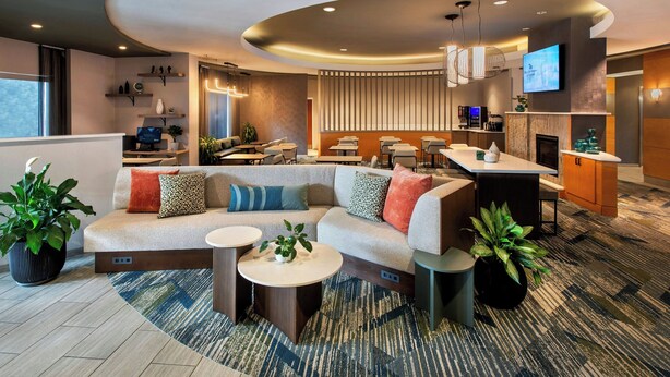 A modern hotel lobby with a curved ceiling, a seating area with sofas and round tables, a kitchen area, and a flat-screen TV mounted on the wall. - SpringHill Suites by Marriott Albany-Colonie (Albany)