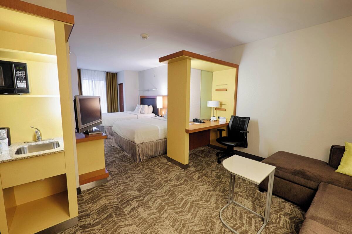 Habitación de hotel con cama, escritorio, silla, mesita, televisor y lavamanos.
