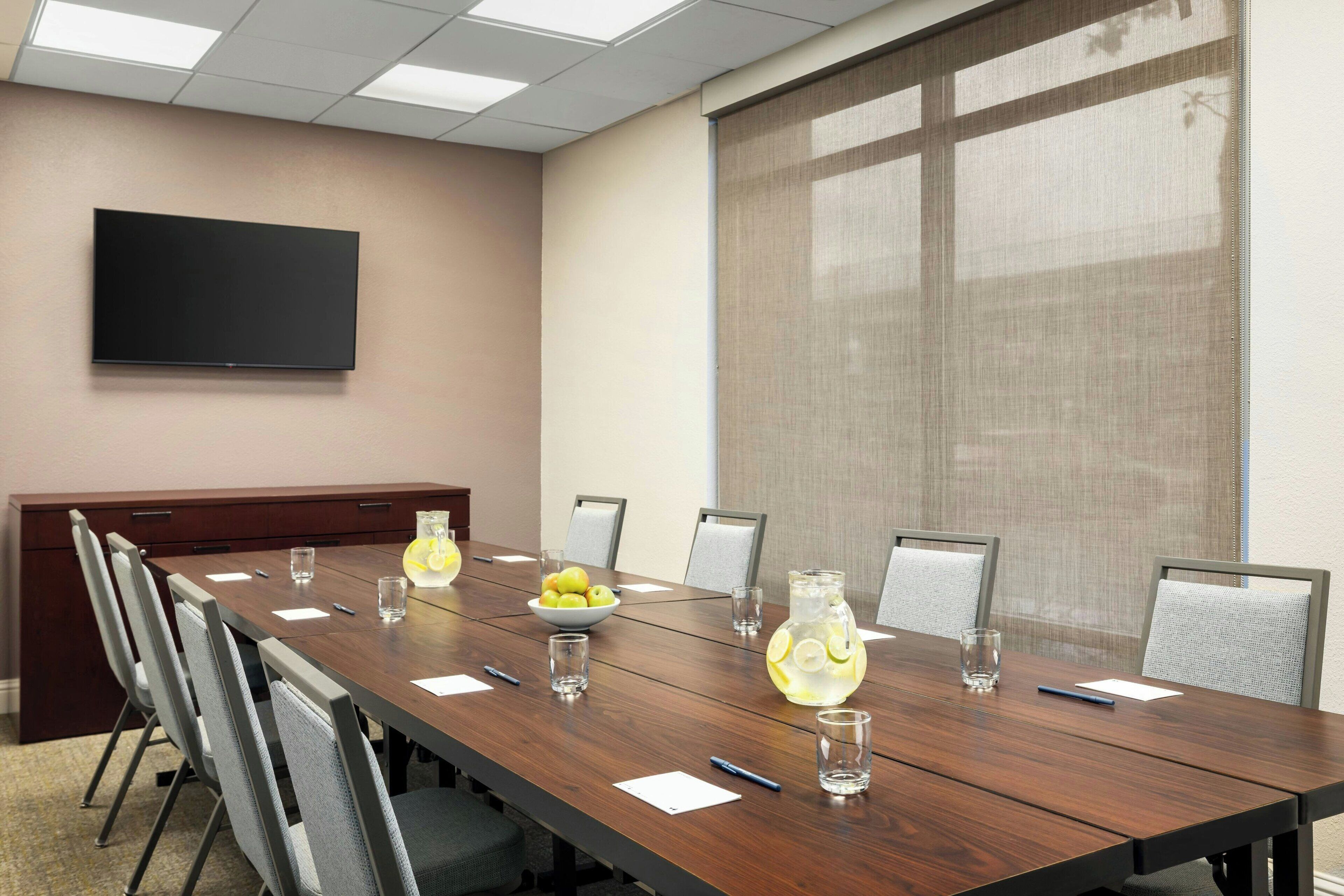A conference room with a long table, chairs, a TV on the wall, and a window with blinds.