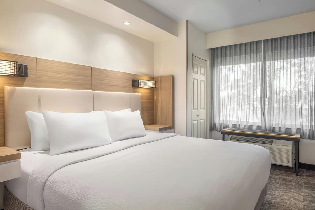 A modern hotel room with a large bed, a bedside table, a desk with a chair, a window with curtains, and a wall-mounted heater.