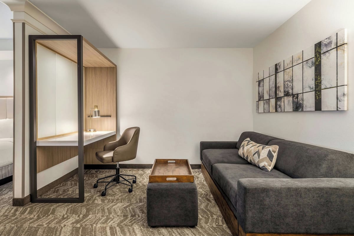 A modern hotel room with a bed, a desk with a lamp, a grey sofa, a small ottoman, and a wall art piece.
