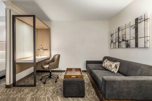 A modern hotel room with a bed, a desk with a lamp, a grey sofa, a small ottoman, and a wall art piece. - SpringHill Suites by Marriott Fresno (Fresno)