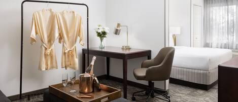 Une chambre d’hôtel comprenant un lit, un bureau avec une lampe, une chaise, une petite table avec une bouteille et des verres, et un porte-vêtements avec deux articles de vêtements.