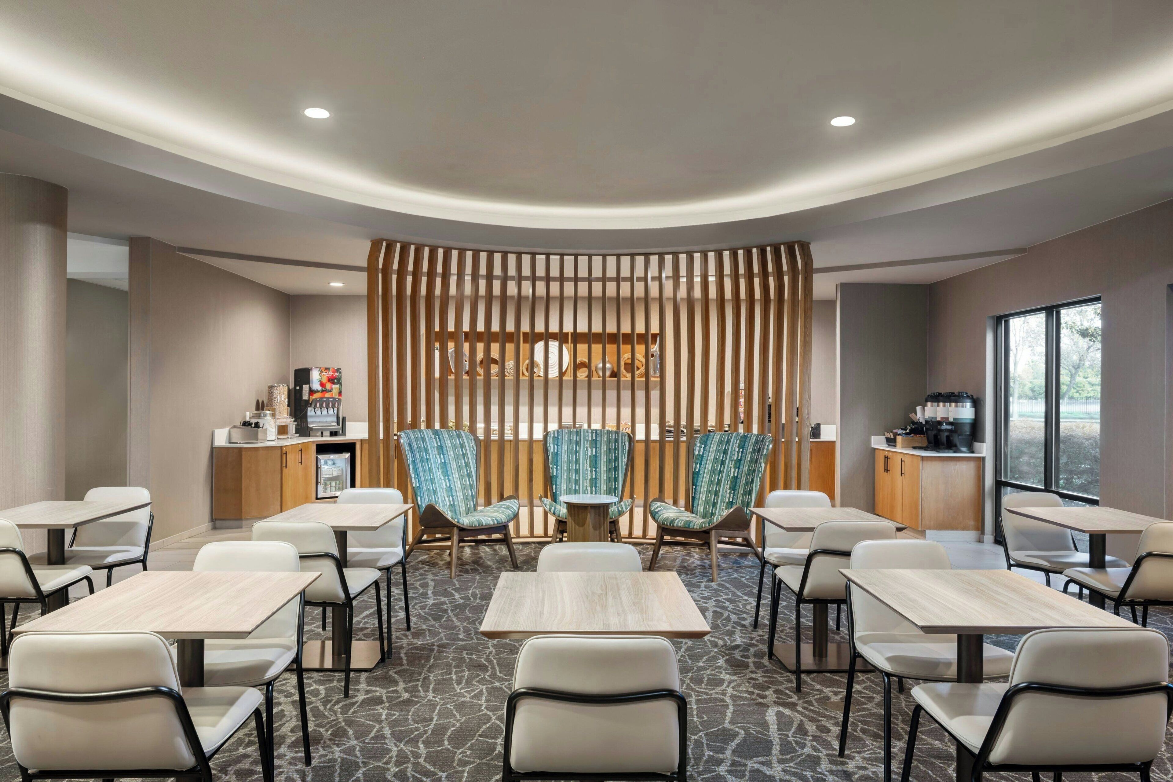 A modern hotel room with a central seating area, a kitchenette, and a large window providing a view.