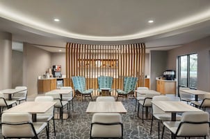 A modern hotel room with a central seating area, a kitchenette, and a large window providing a view.