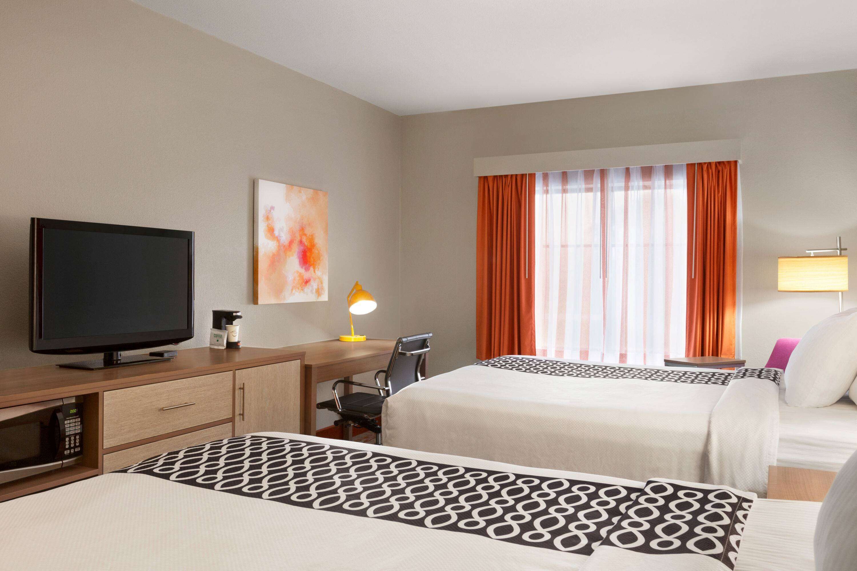 A hotel room with two beds, a desk with a TV, a desk lamp, and a window with orange curtains.