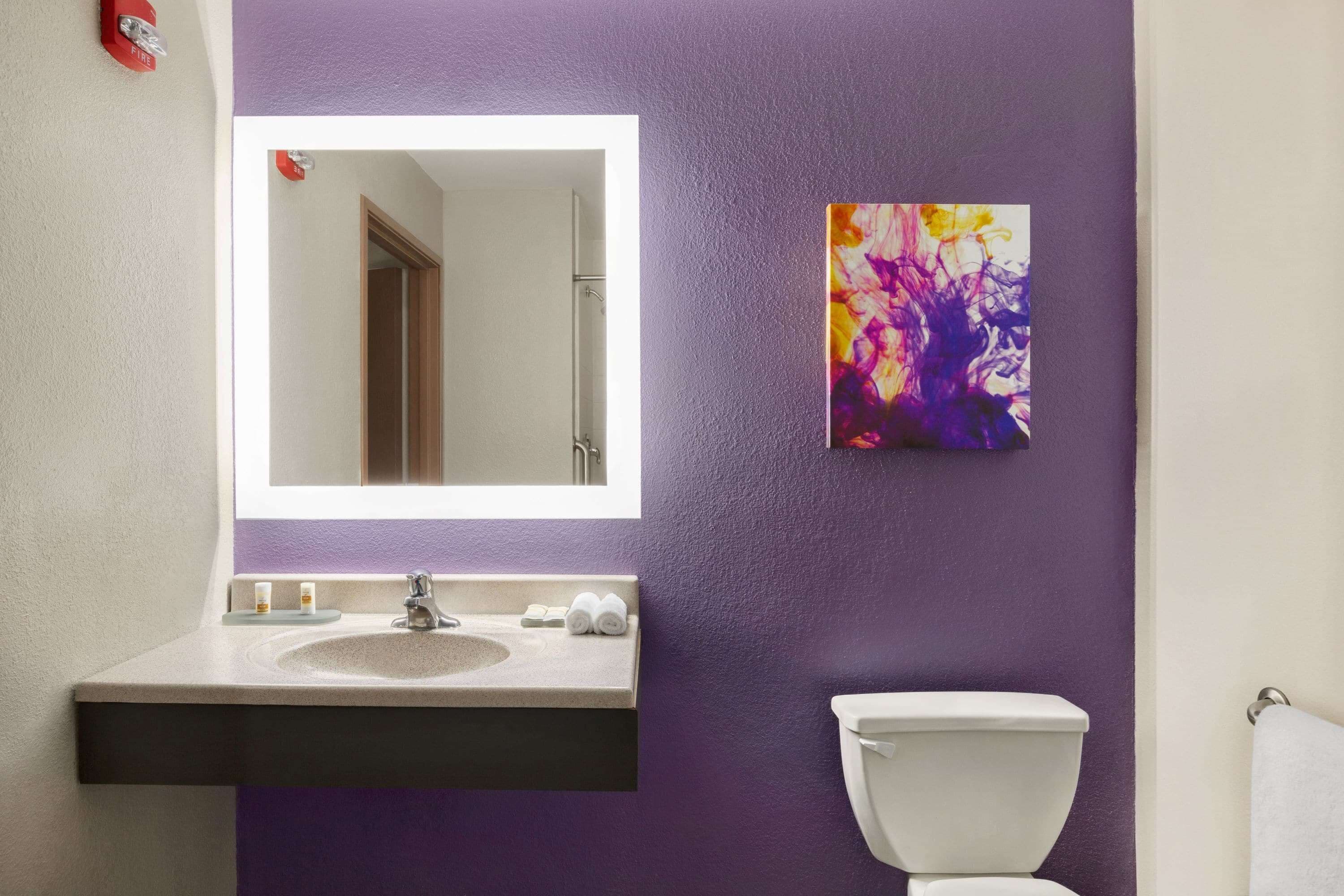 A bathroom with a purple wall, a white sink, a mirror, a toilet, and a colorful abstract painting.