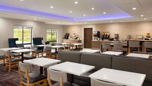 A modern restaurant with white tables and grey cushioned booths, a kitchen area, and large windows. - La Quinta Inn & Suites by Wyndham Lackawanna (Buffalo)