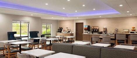 A modern restaurant with white tables and grey cushioned booths, a kitchen area, and large windows.