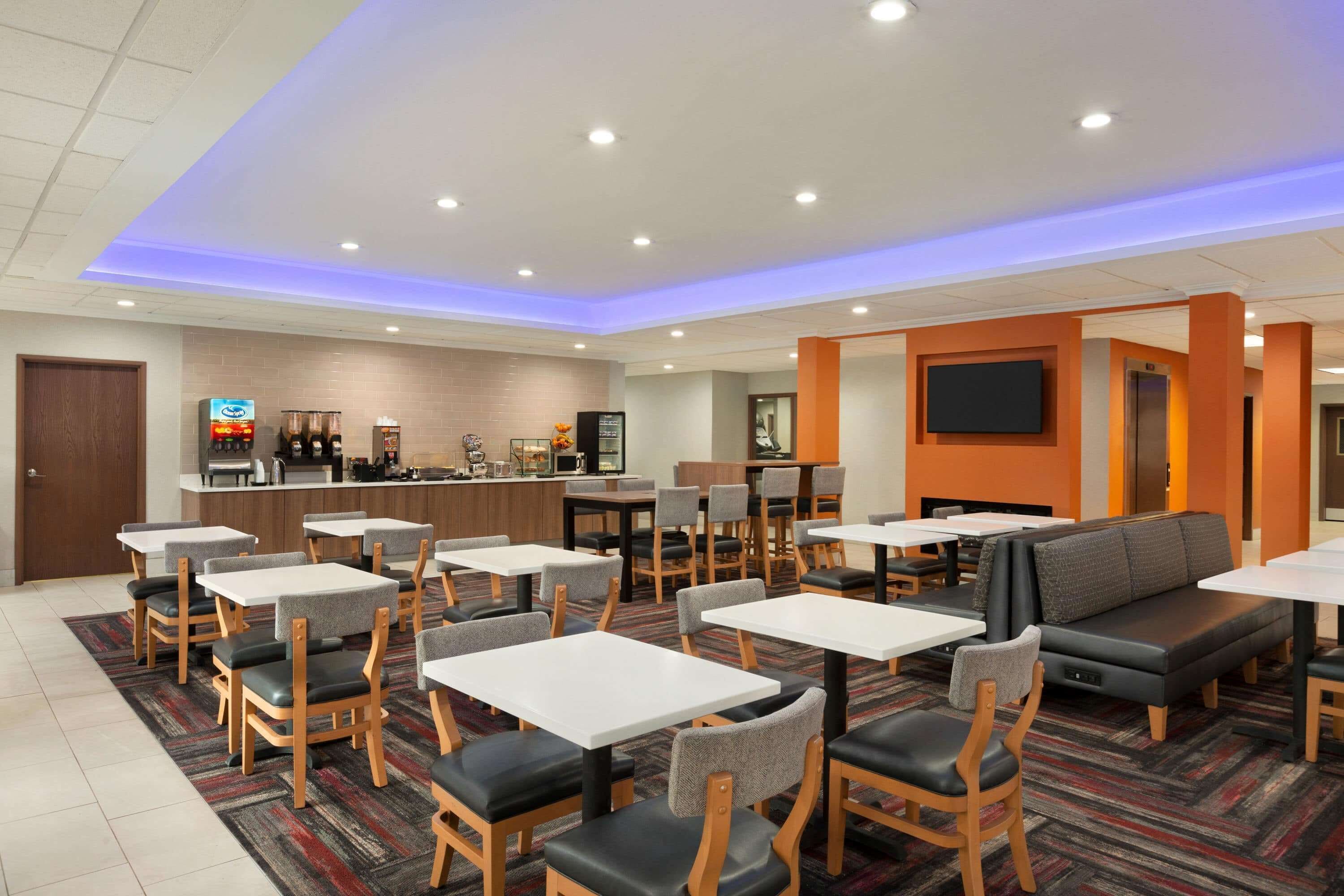 Eine Hotelhalle mit mehreren Sitzbereichen, darunter Tische und Stühle, eine Rezeption mit einer Kaffeemaschine und ein an der Wand befestigtes Fernsehgerät.
