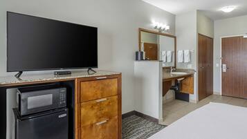 Quarto de hotel com TV de tela plana, micro-ondas, geladeira, armário de madeira e banheiro com pia, espelho e porta-toalhas.