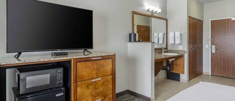 A hotel room with a flat-screen TV, a microwave, a refrigerator, a wooden cabinet, a bathroom with a sink, mirror, and towel rack.