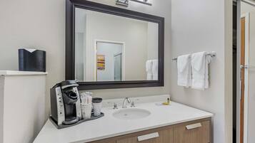 Un baño con lavamanos blanco, cafetera, espejo y perchero para toallas.