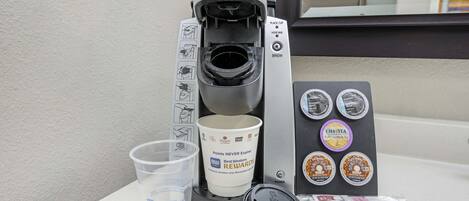 Una cafetera Keurig con una taza de café, un vaso reutilizable y un soporte para cápsulas con diversas opciones de café.