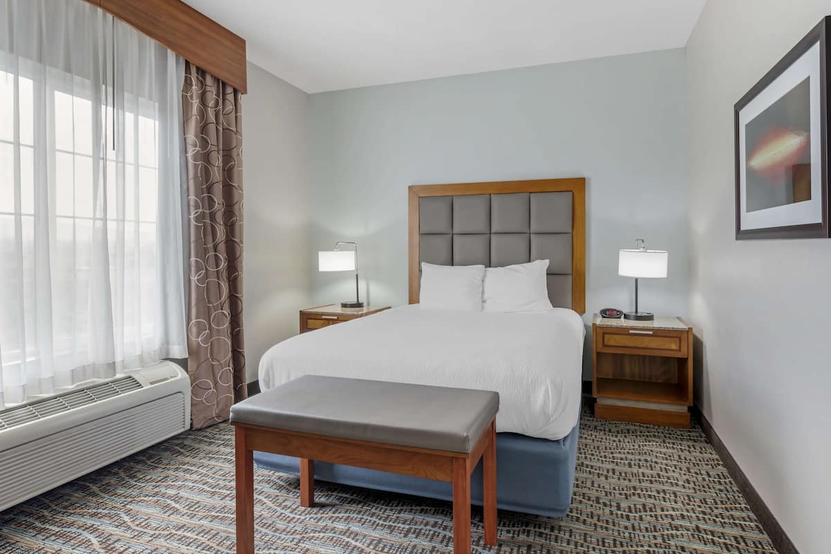 A hotel room with a bed, two bedside tables with lamps, a bench, a window with curtains, and a wall-mounted picture.
